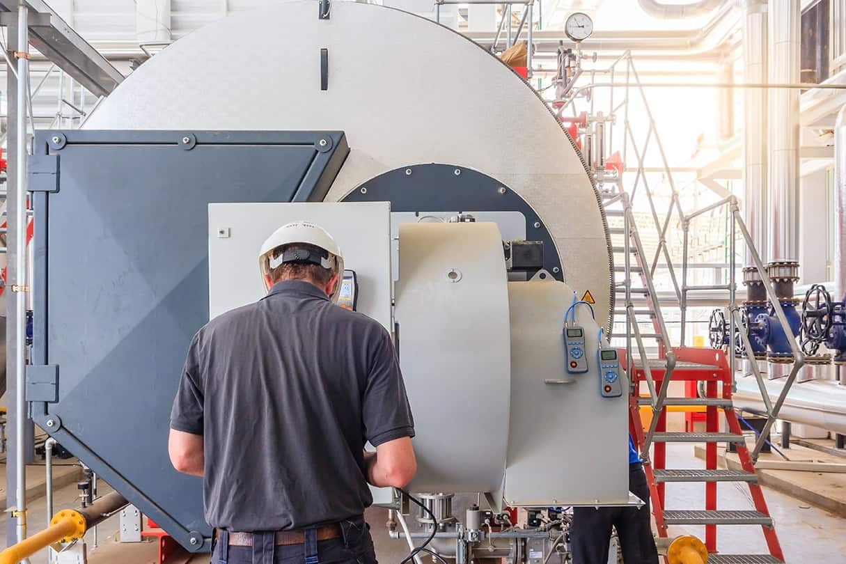 A boiler technician, with his back to the camera, inspecting the controls on a commercial boiler in a boiler room.